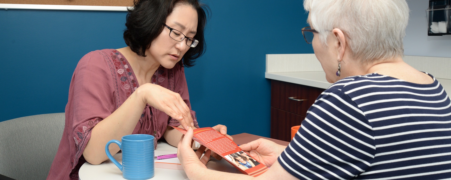 An Asian PMHNP providing education to an elderly female patient in a break room setting.
